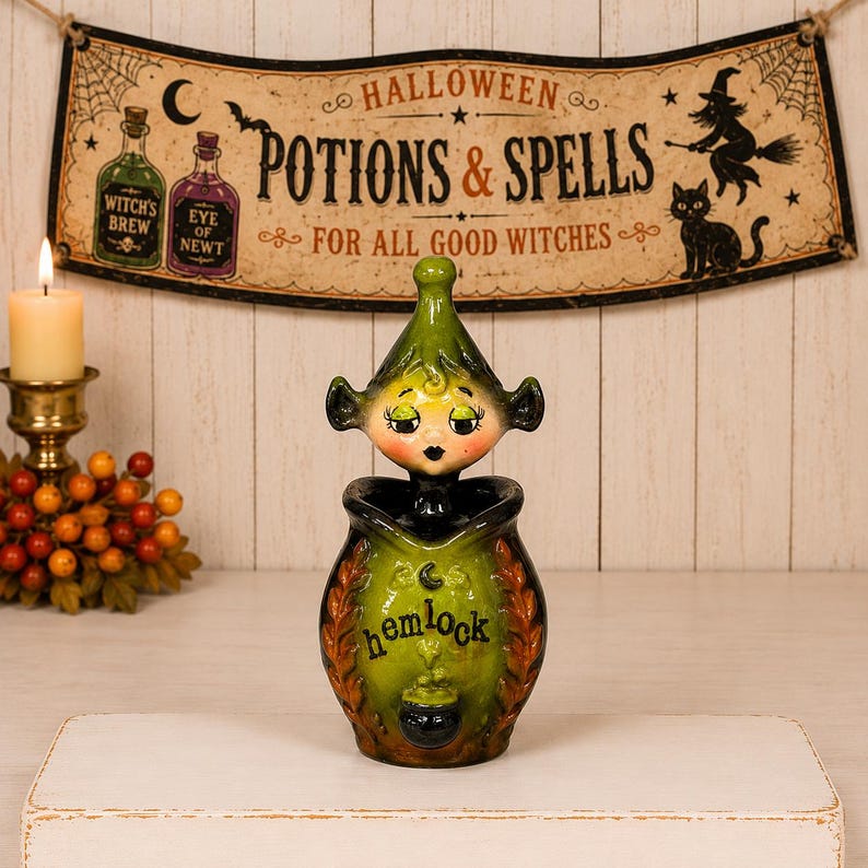 May include: A ceramic jar featuring a green, pointed-hat figure with closed eyes and a black-painted face. The jar body is green with orange leaf accents, a black crescent moon, and the word "hemlock" printed in black above a small cauldron graphic.