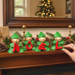 May include: Four ceramic Christmas elf figurines in green and red, spelling "NOEL". Each elf has a green hat and body, holding a red letter. The figurines are on a wooden surface with a Christmas tree in the background.