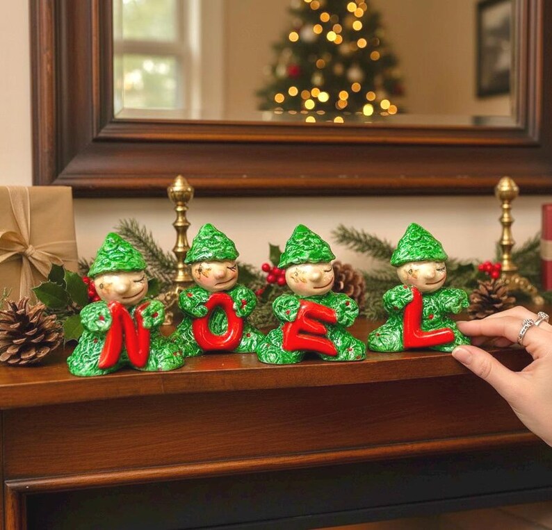 May include: Four ceramic Christmas elf figurines in green and red, spelling "NOEL". Each elf has a green hat and body, holding a red letter. The figurines are on a wooden surface with a Christmas tree in the background.
