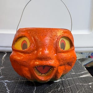 May include: Vintage orange pumpkin-shaped Halloween candy pail with a smiling face and a metal handle. The pail has yellow eyes with red accents and a jagged mouth. The textured surface gives it a classic, aged appearance.