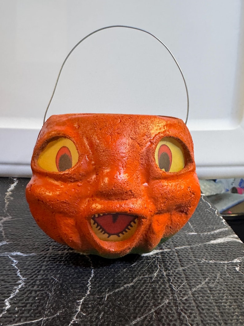 May include: Vintage orange pumpkin-shaped Halloween candy pail with a smiling face and a metal handle. The pail has yellow eyes with red accents and a jagged mouth. The textured surface gives it a classic, aged appearance.