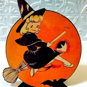 May include: A vintage Halloween decoration featuring a witch flying on a broomstick in front of a jack-o'-lantern moon. The witch is wearing a black dress and a pointed hat with white stripes. The moon has a smiling face with a single eye.