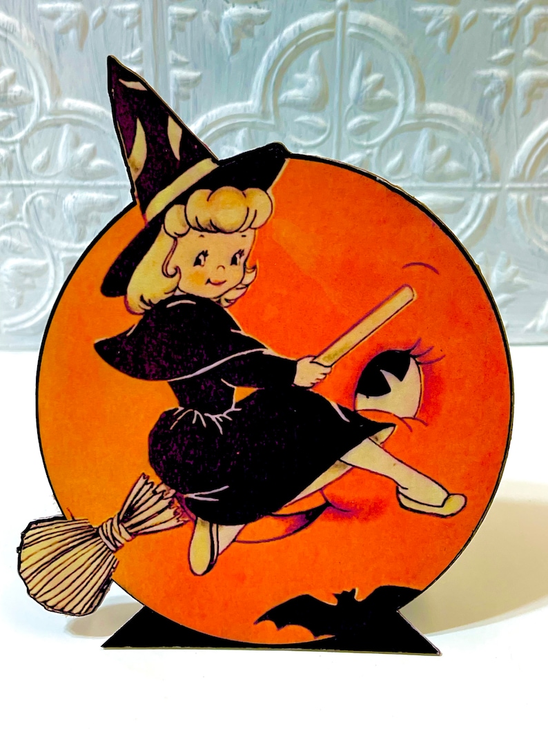May include: A vintage Halloween decoration featuring a witch flying on a broomstick in front of a jack-o'-lantern moon. The witch is wearing a black dress and a pointed hat with white stripes. The moon has a smiling face with a single eye.