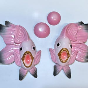May include: Two pink and white ceramic fish figurines with open mouths and gold accents, alongside three pink pearl-like spheres. The fish have black-tipped fins and tails. The items are likely decorative.