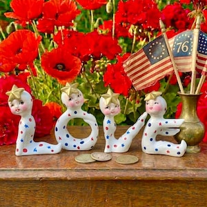 May include: A decorative arrangement featuring the word "LOVE" spelled out with whimsical ceramic figures. Each figure is white with red and blue accents, a star-shaped headpiece, and a smiling face. The figures are arranged on a wooden surface with coins and a vase with small American flags.