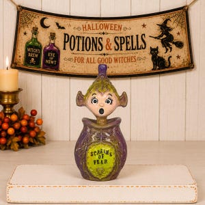 May include: A decorative ceramic figurine featuring a purple and green potion bottle base with the text "SCREAMS OF FEAR" and a sculpted head with wide eyes and an open mouth emerging from the top, set against a Halloween-themed wall sign.