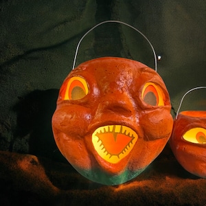 May include: Orange Halloween pumpkin bucket with a smiling face, glowing eyes, and a jagged-tooth grin. The bucket has a metal handle and a green base. The pumpkin is lit from within, creating a spooky effect.