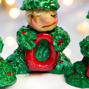 May include: A festive Christmas ornament depicting a figure in a green leafy outfit holding a red letter "O". The figure has a painted face with closed eyes and a sad expression. The ornament is hand-painted with a glossy finish.