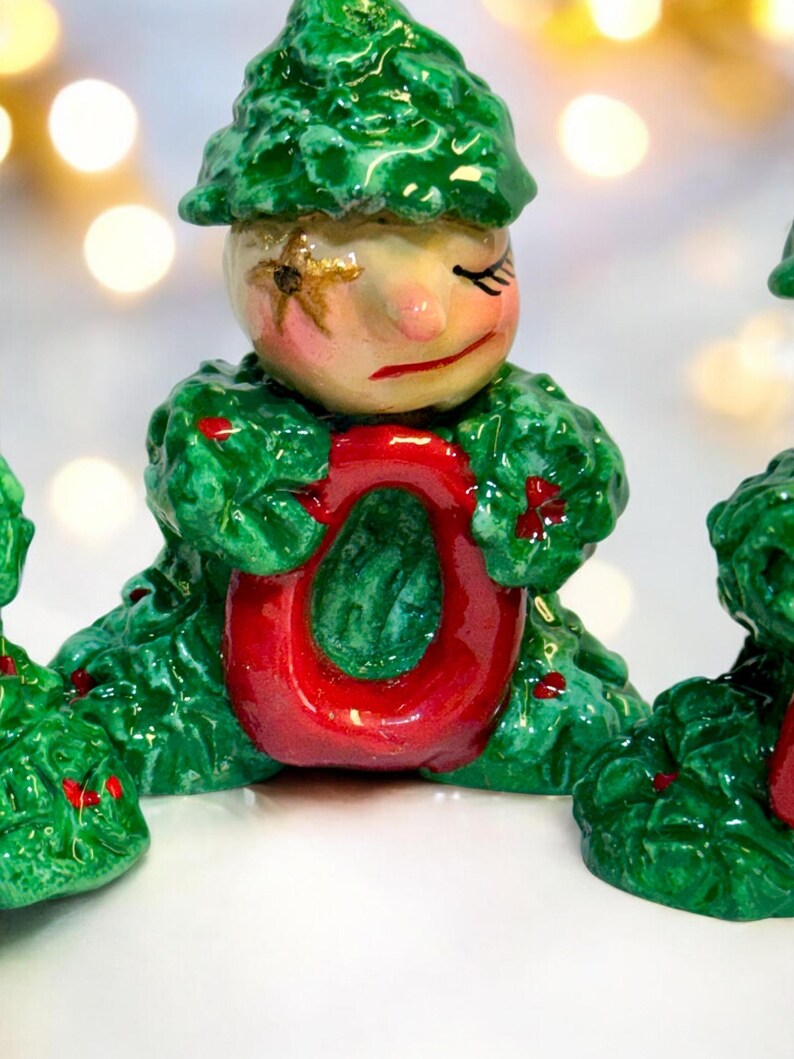 May include: A festive Christmas ornament depicting a figure in a green leafy outfit holding a red letter "O". The figure has a painted face with closed eyes and a sad expression. The ornament is hand-painted with a glossy finish.
