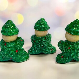 May include: Three decorative figurines with a green, textured body and a light beige head. The figurines have red accents and a glossy finish. They are arranged on a white surface with a blurred background.