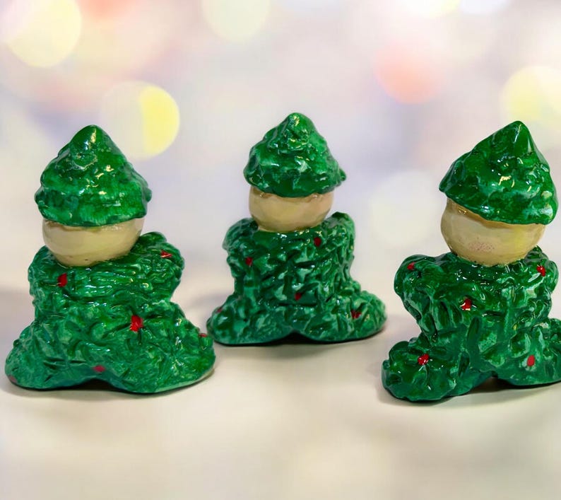 May include: Three decorative figurines with a green, textured body and a light beige head. The figurines have red accents and a glossy finish. They are arranged on a white surface with a blurred background.