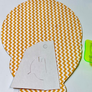 May include: A white paper template with a cut-out design for a stand-up figure. The template is placed on top of a yellow and white chevron patterned paper circle.