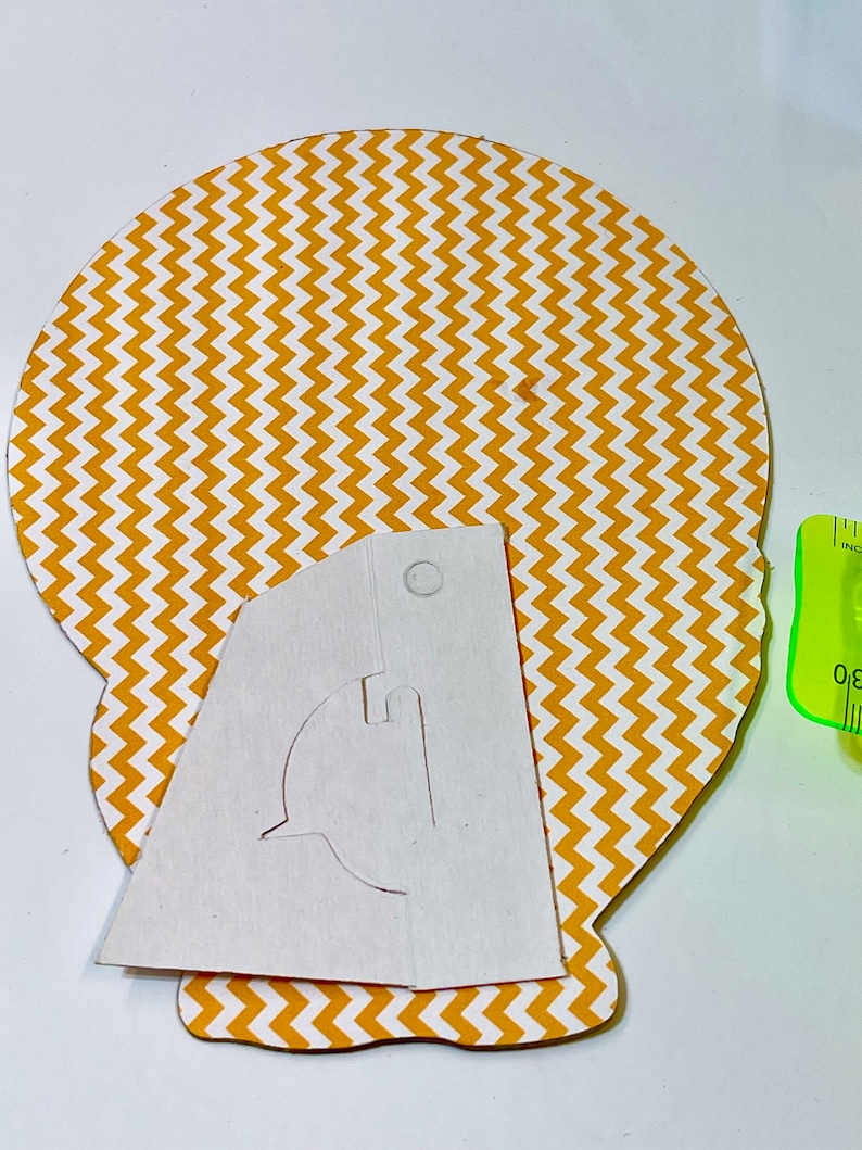 May include: A white paper template with a cut-out design for a stand-up figure. The template is placed on top of a yellow and white chevron patterned paper circle.