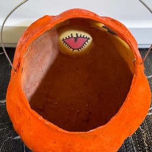 May include: Orange, pumpkin-shaped Halloween candy pail with a metal handle. The inside of the pail is brown. A small, oval-shaped image of a monster's eye is visible inside the pail.