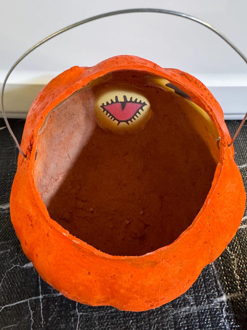 May include: Orange, pumpkin-shaped Halloween candy pail with a metal handle. The inside of the pail is brown. A small, oval-shaped image of a monster's eye is visible inside the pail.