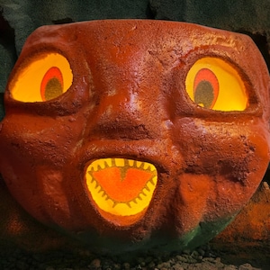May include: A large, orange, carved pumpkin with glowing yellow eyes and mouth. The pumpkin has a textured surface and a friendly, cartoonish expression. The interior of the eyes and mouth glow with a warm, inviting light, suggesting a Halloween theme.