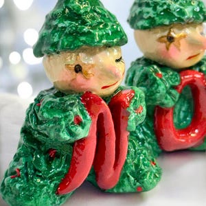 May include: Two ceramic elf figurines with green textured hats and coats. The elves have pale skin, rosy cheeks, and red lips. They hold a bright red, ribbon-like object. The background is blurred with bokeh lights.