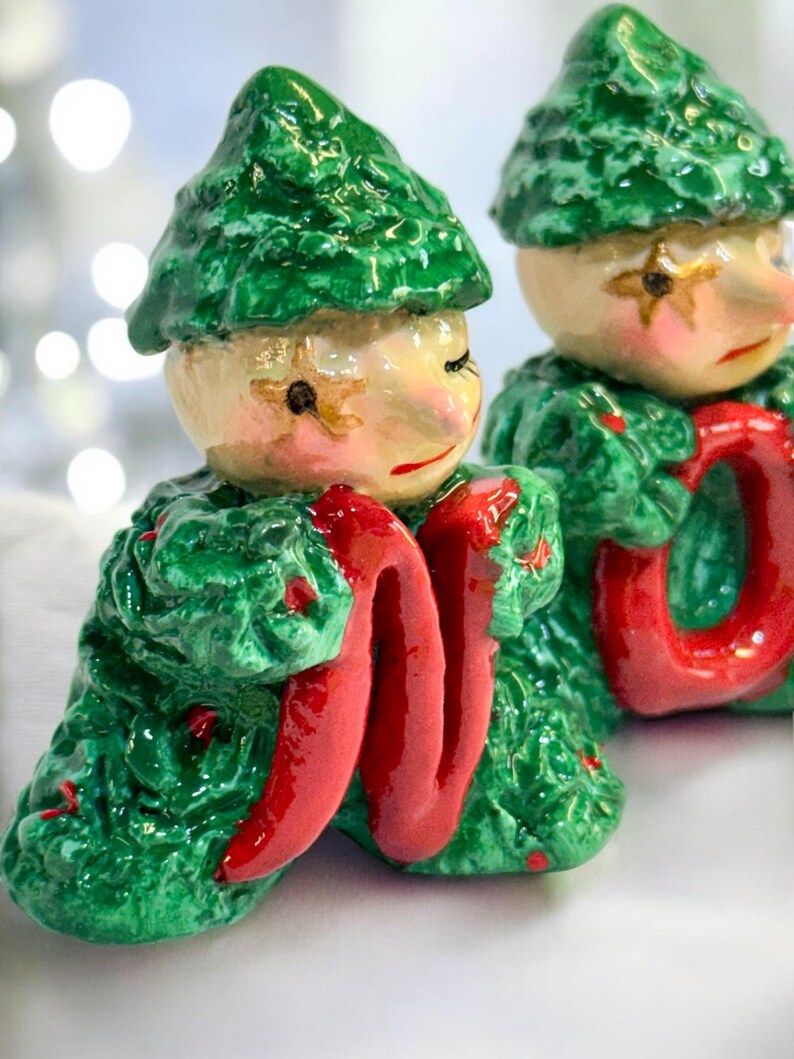 May include: Two ceramic elf figurines with green textured hats and coats. The elves have pale skin, rosy cheeks, and red lips. They hold a bright red, ribbon-like object. The background is blurred with bokeh lights.