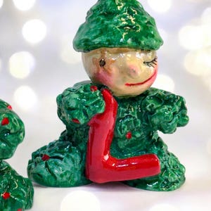 May include: A ceramic elf figurine, predominantly green with red details. The elf is holding a red letter "L". The figurine has a smiling face with closed eyes, and is wearing a pointed hat. The figurine is likely handmade.
