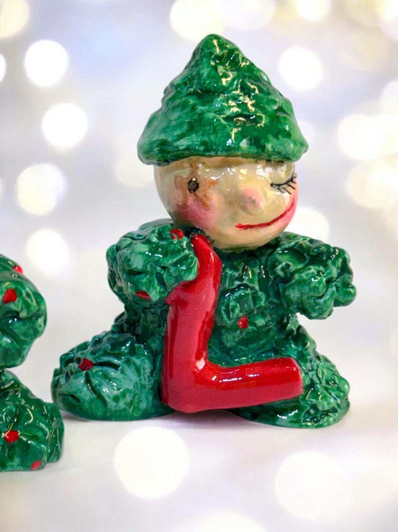 May include: A ceramic elf figurine, predominantly green with red details. The elf is holding a red letter "L". The figurine has a smiling face with closed eyes, and is wearing a pointed hat. The figurine is likely handmade.
