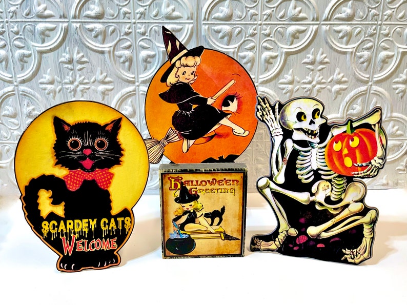 May include: Four vintage Halloween decorations: a black cat with a red bow tie, a witch flying on a broomstick, a skeleton holding a pumpkin, and a wooden block with a witch and the text "Halloween Greeting".