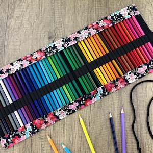 May include: A colorful pencil roll with a floral design. The roll is filled with a variety of colored pencils, arranged in a rainbow spectrum. The fabric has a black background with pink and white flowers. A few pencils are scattered around the roll.