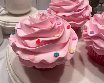 May include: Three decorative cupcakes featuring swirls of pink frosting topped with small, flat, multi-colored circular sprinkles. Each cupcake sits in a dark pink paper liner, arranged on a round, off-white, distressed wooden pedestal stand.