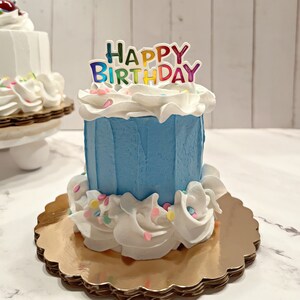 Pu&ograve; includere: Una piccola torta di compleanno blu con glassa bianca e codette colorate. La torta ha un topper "Happy Birthday" con lettere arcobaleno.