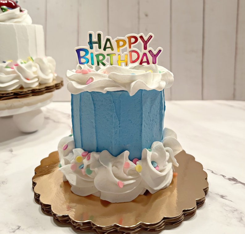 Pu&ograve; includere: Una piccola torta di compleanno blu con glassa bianca e codette colorate. La torta ha un topper "Happy Birthday" con lettere arcobaleno.