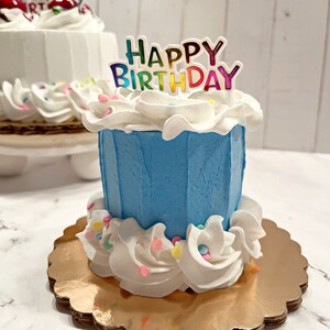 Pu&ograve; includere: Una piccola torta di compleanno blu con glassa bianca e codette colorate. La torta &egrave; decorata con un topper "Happy Birthday" con lettere arcobaleno.