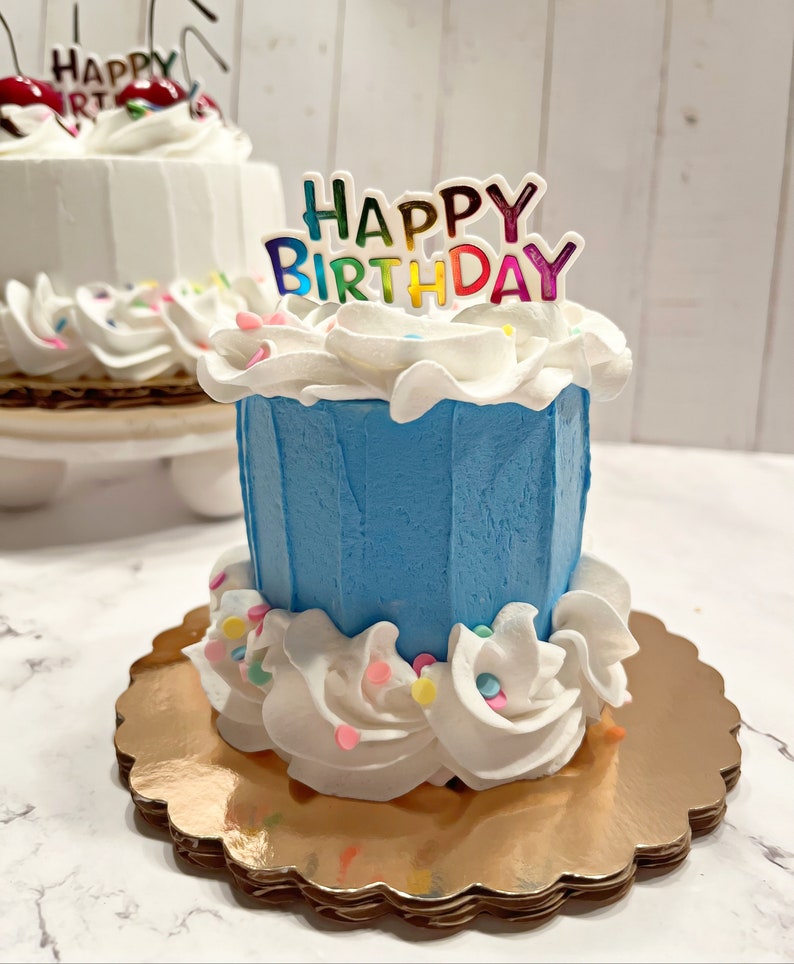 Pu&ograve; includere: Una piccola torta di compleanno blu con glassa bianca e codette colorate. La torta &egrave; decorata con un topper "Happy Birthday" con lettere arcobaleno.