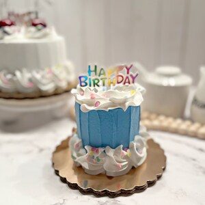 Pu&ograve; includere: Una piccola torta di compleanno blu con glassa bianca e codette colorate. La torta &egrave; decorata con un topper "Happy Birthday" con lettere arcobaleno.