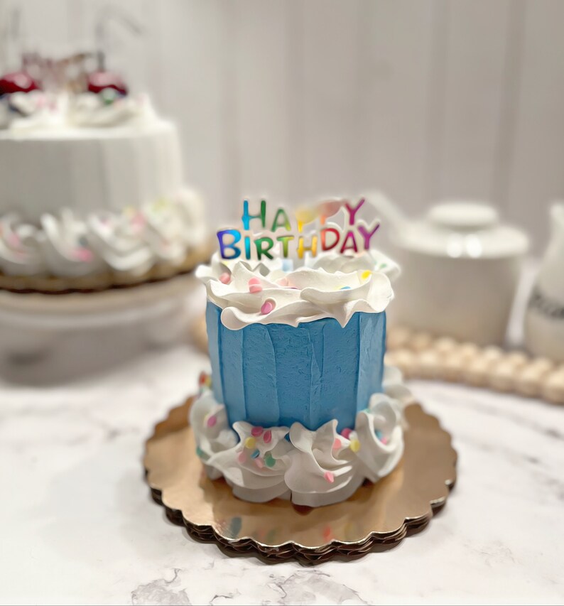 Pu&ograve; includere: Una piccola torta di compleanno blu con glassa bianca e codette colorate. La torta &egrave; decorata con un topper "Happy Birthday" con lettere arcobaleno.