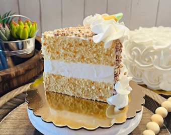 May include: A slice of white cake with a layer of frosting and a sprinkle of brown crumbs. The cake is on a gold cake stand with a white base. The cake is decorated with white frosting and a small piece of orange candy.
