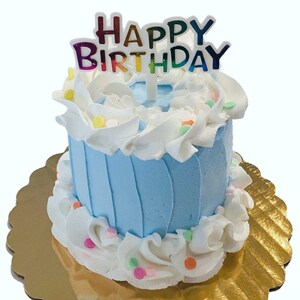 Pu&ograve; includere: Una piccola torta di compleanno blu e bianca con un topper "Buon compleanno". La torta &egrave; decorata con glassa bianca e codette colorate.