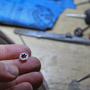 May include: A silver-colored star-shaped charm with a cut-out design. The charm is being held in a person's hand.