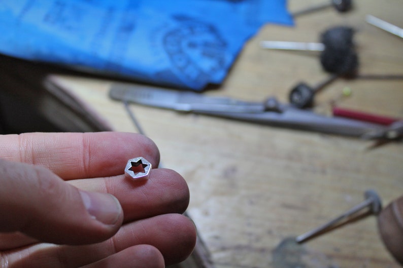May include: A silver-colored star-shaped charm with a cut-out design. The charm is being held in a person's hand.