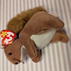 Puede incluir: Un juguete de peluche de ardilla marrón con una cola esponjosa y un vientre blanco. El juguete tiene una etiqueta roja en forma de corazón con una estrella dorada y la palabra "Ty". La ardilla está sobre una superficie rayada.