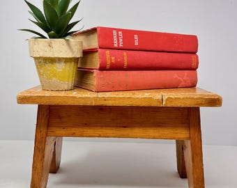 May include: A small, wooden, rectangular stool with four legs. Three red books are stacked on top, with the titles visible. A small, yellow and beige pot with a green plant sits on the left side of the stool.