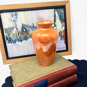 Op de afbeelding: Een glanzende oranje keramische vaas staat op twee antieke boeken met rode covers en blauwe labels. Een ingelijst schilderij van een winterlandschap staat op de achtergrond. De vaas heeft een afgeronde body en een uitlopende rand.