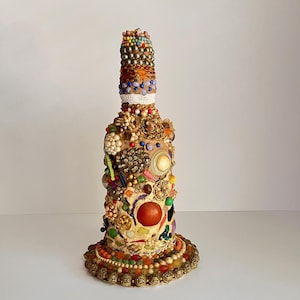 Peut inclure: Une bouteille en verre entièrement recouverte d'une mosaïque de perles colorées, de boutons et d'autres bibelots. La bouteille a une base dorée et un sommet arrondi.