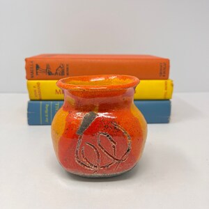 Puede incluir: Un pequeño jarrón de cerámica naranja y amarillo con un diseño negro. El jarrón está sobre una pila de libros.