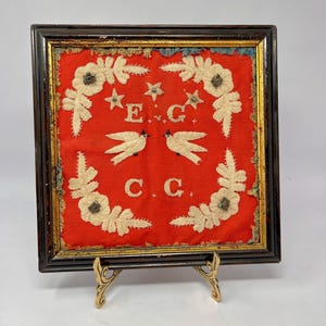May include: Framed vintage needlepoint with a red background. The design features two white birds, floral accents, stars, and the letters "E.G." and "C.C." in white. The frame is dark brown with gold trim, and the piece is displayed on a decorative stand.