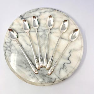 May include: Six silver spoons arranged on a round marble serving tray. The spoons have a classic design with a decorative pattern on the handle. The tray has a white and gray marble pattern.