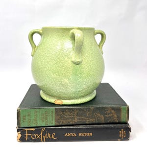 May include: A light green ceramic vase with two handles, sitting atop two vintage books. The vase has a speckled texture and a rounded shape. The books are dark green and black, with gold lettering.