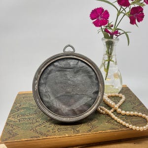 May include: A round, silver-toned picture frame with a dark interior sits atop a stack of antique books. A small glass vase holds vibrant pink flowers. A strand of pearls is draped nearby, creating a vintage aesthetic.