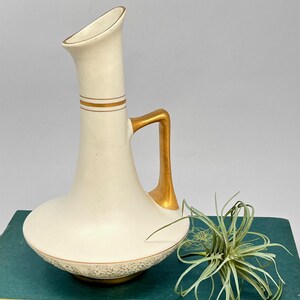 Può includere: Brocca vintage color crema con manico e finiture dorate. La brocca ha una base bulbosa e un collo sottile con apertura svasata. Due fasce dorate circondano il collo. Un motivo floreale decorativo adorna la base.