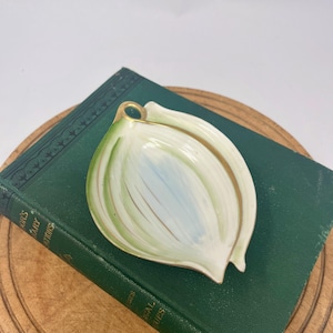 以下が含まれることがあります： 金色の縁と取っ手が付いた、玉ねぎ型の陶器の皿。皿は、緑と青のアクセントが付いた白いベースが特徴です。ヴィンテージの緑色の本の上に置かれています。