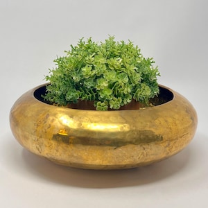 Puede incluir: Un cuenco bajo y ancho de latón martillado con un acabado dorado pulido. El cuenco contiene una pequeña y densa planta artificial verde con follaje texturizado similar a un helecho, situada en el centro de la abertura circular del recipiente.