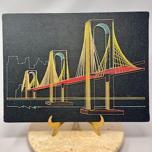 May include: A string art depiction of a suspension bridge on a black background. The bridge features yellow support cables, a red roadway, and blue arches. The artwork is displayed on a marble stand with gold-colored supports.